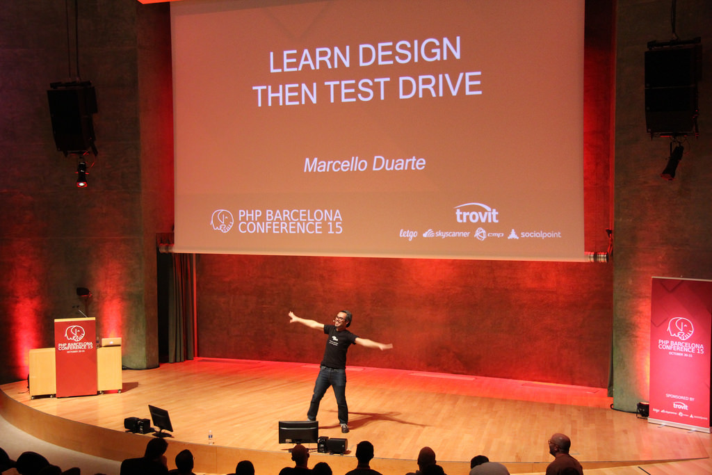 Marcello Duarte on stage at PHP Barcelona Conference 2015, arms outstretched under the slide 'Learn Design Then Test Drive'